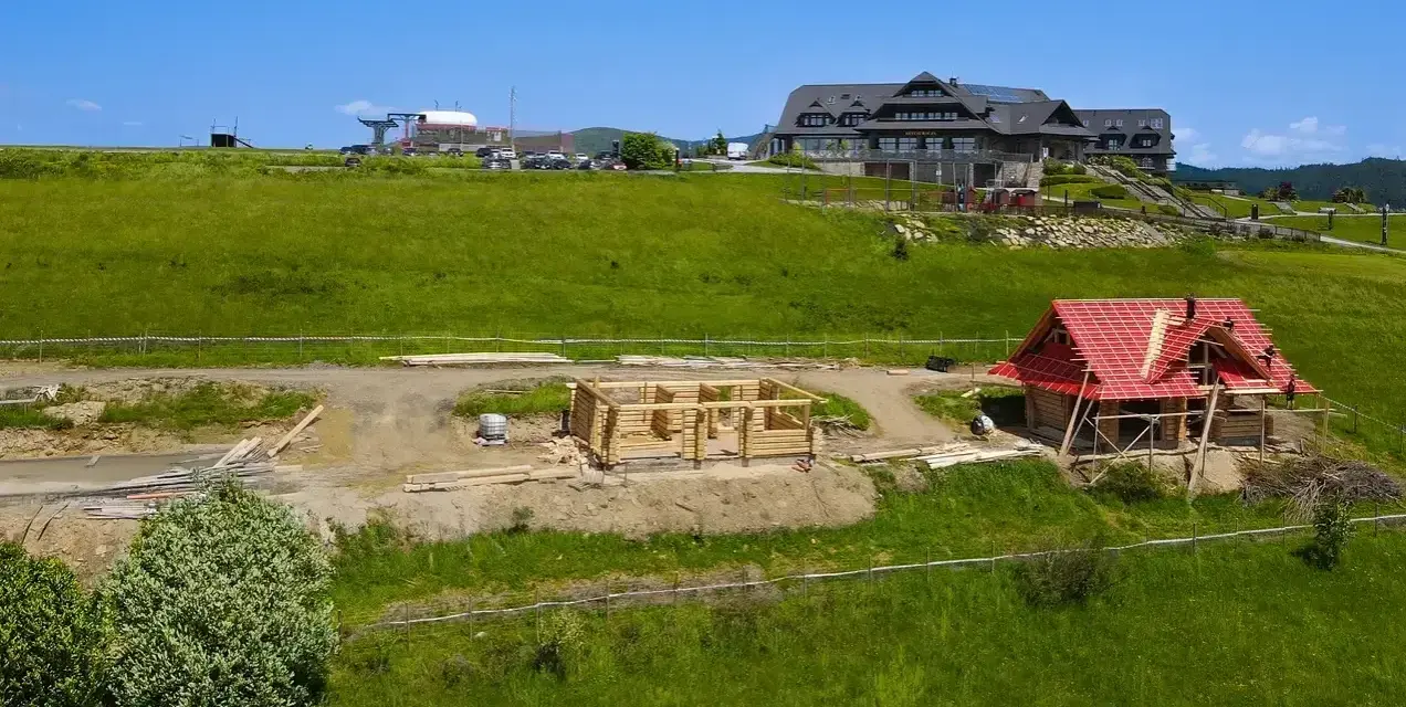 Budowa domu z bali w górach. Widać fundamenty i ściany z bali, a także dom z czerwonym dachem w trakcie prac. To początek budowy, jak wyczyścić dom z bali, by służył latami.