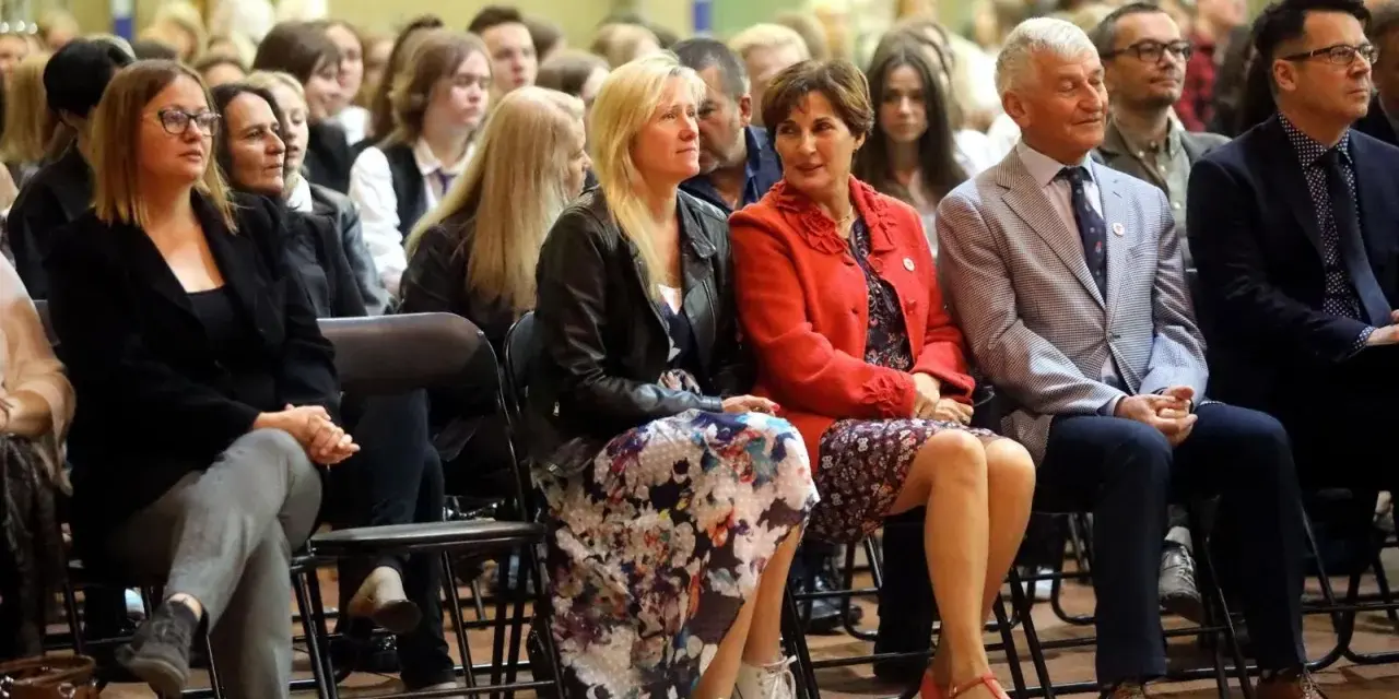 Uczestnicy siedzą na krzesłach, słuchając uważnie. Wśród nich widać osoby, które mogą być na radzie pedagogicznej, w tym kobiety w eleganckich strojach i mężczyzn w garniturach.