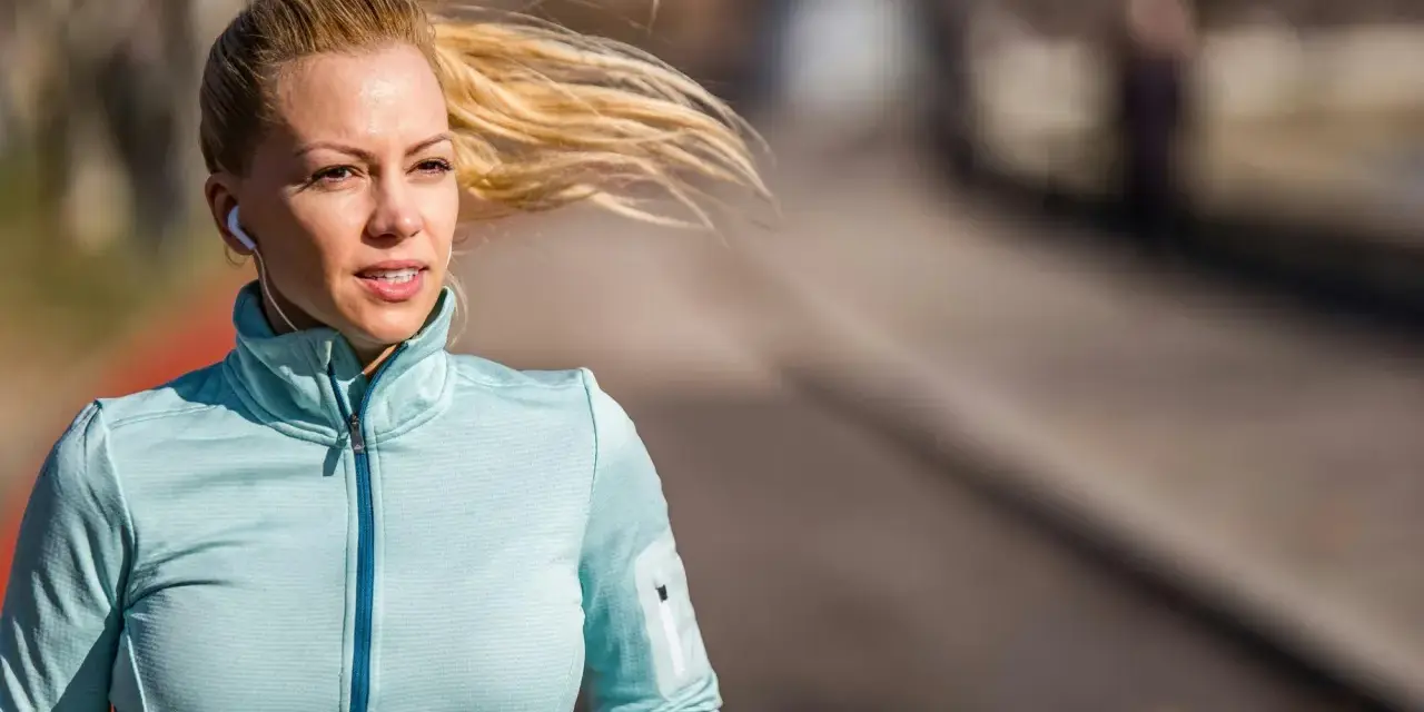Kvinna med blont hår i hästsvans springer utomhus, lyssnar på musik med hörlurar. Hon är fokuserad på att springa 5 km tid.