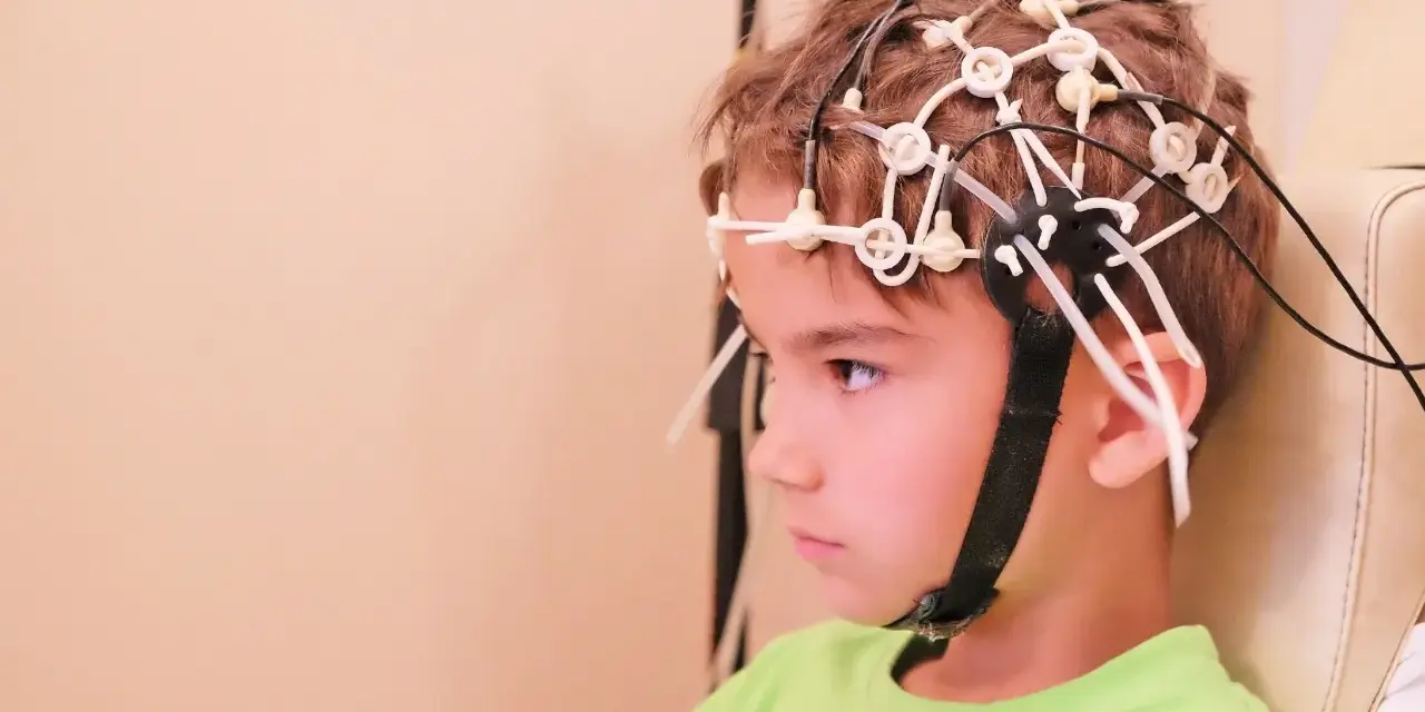 Chłopiec w zielonej koszulce ma na głowie czepki do eeg głowy, z przewodami podłączonymi do elektroencefalografu.