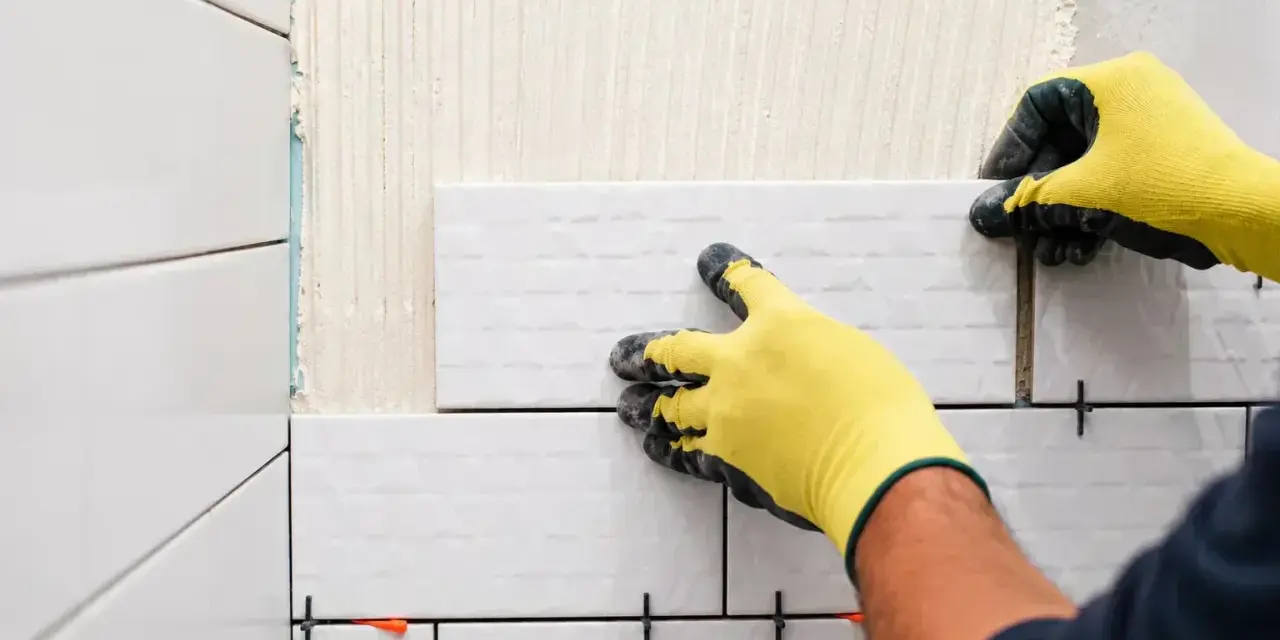 Pose-t-on coller du carrelage avec du ciment ? Oui, un carreleur pose du carrelage blanc sur un mur avec des croisillons.