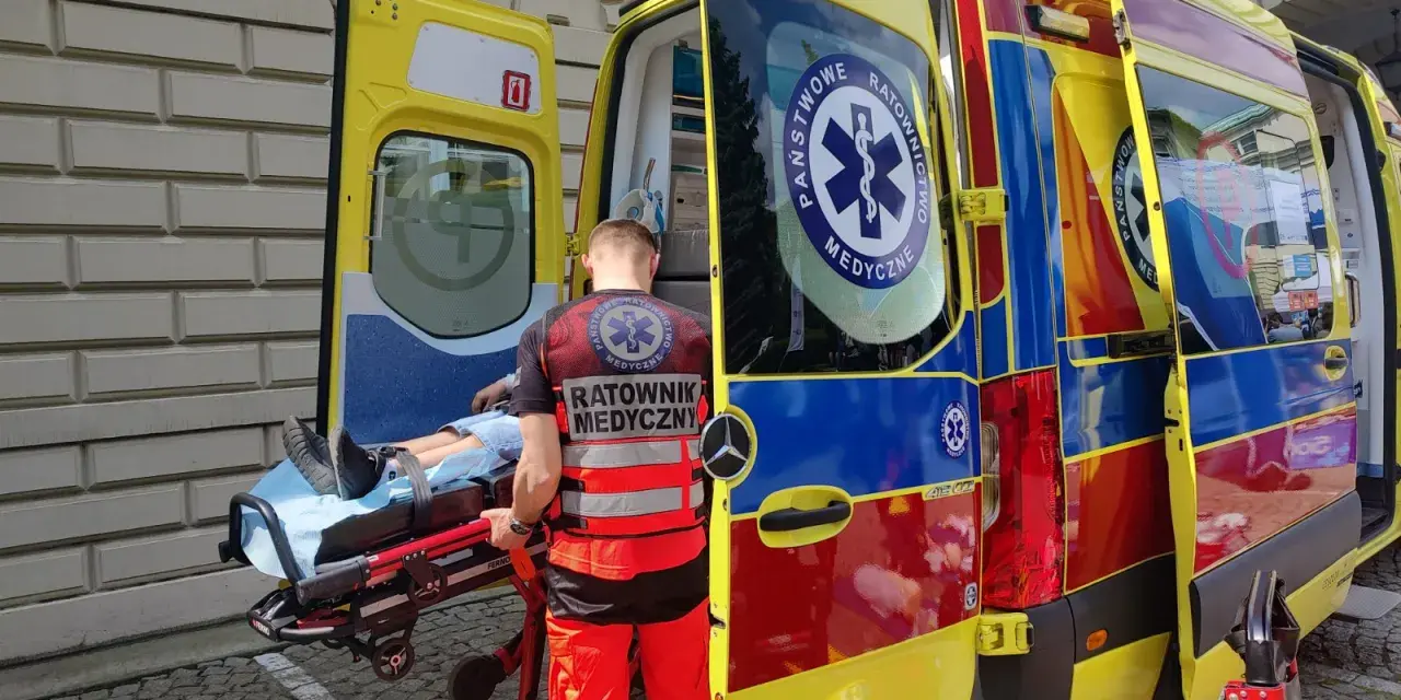 Ratownik medyczny ładuje pacjenta na noszach do karetki. Na jego kamizelce widnieje napis "RATOWNIK MEDYCZNY".