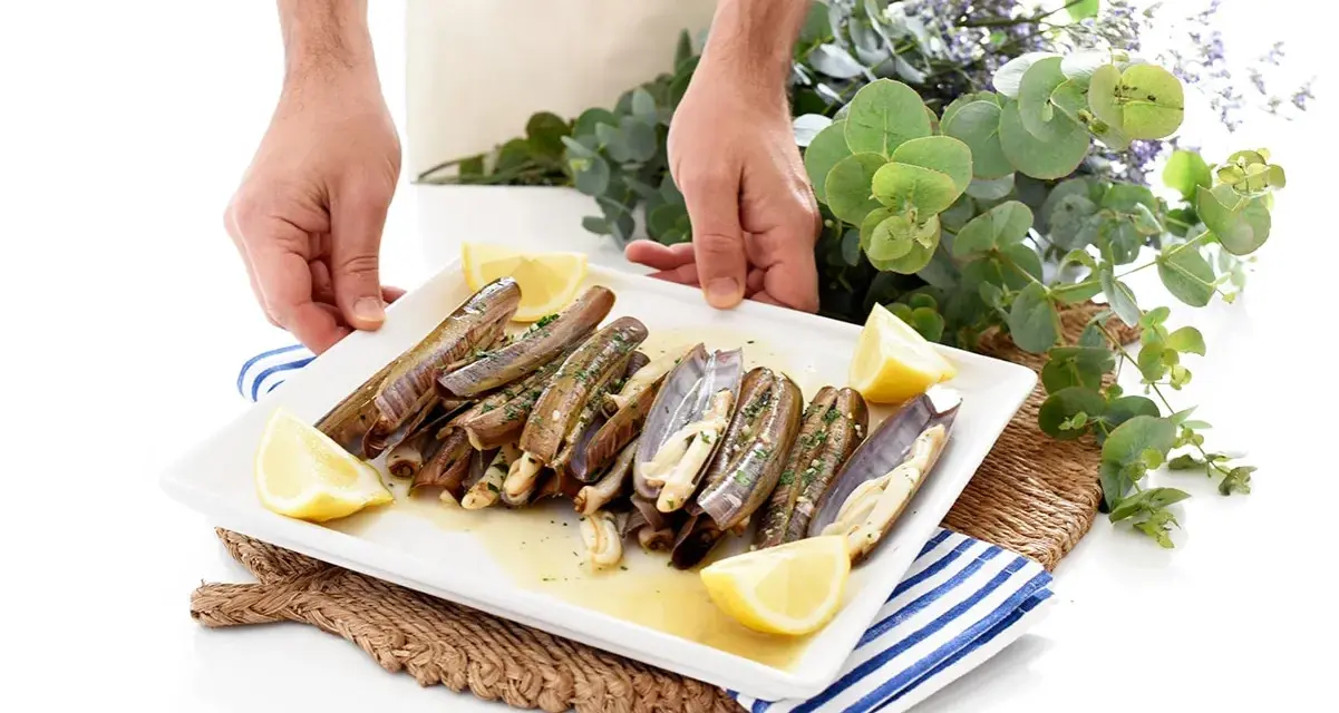 Un plato blanco con navajas a la plancha, limón y perejil. Unas manos sostienen el plato, decorado con eucalipto y flores moradas.