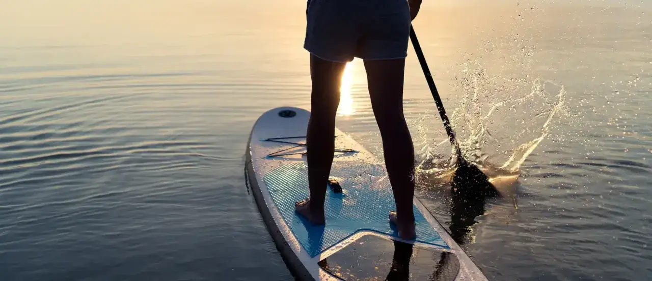 Person lernt SUP auf ruhigem Wasser bei Sonnenuntergang. Das Paddel spritzt Wasser auf.
