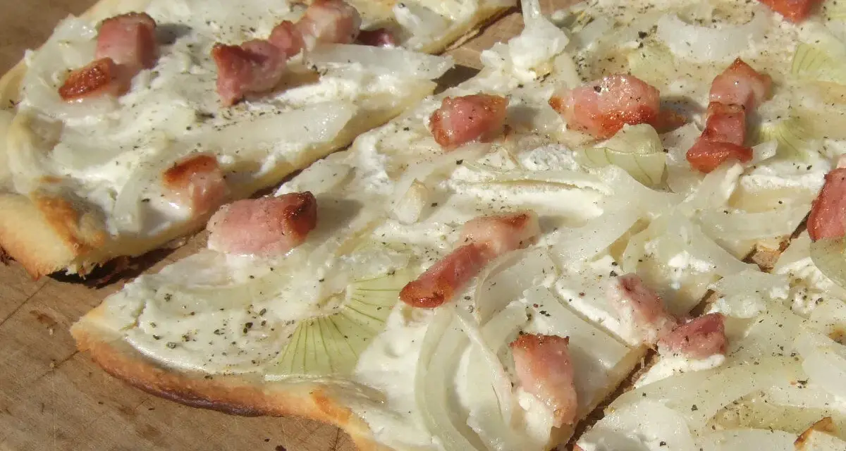 Flammkuchen mit dünnen Zwiebelringen und Speckwürfeln auf Crème fraîche. Ein köstlicher Belag für den knusprigen Teig.