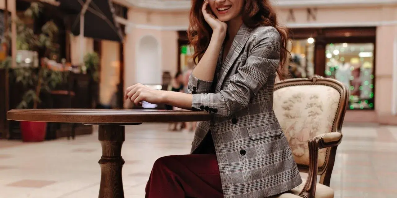 Elegancka kobieta w okularach i marynarce w kratę, prezentująca stylizacje z bordowymi spodniami, siedzi przy stoliku w kawiarni.