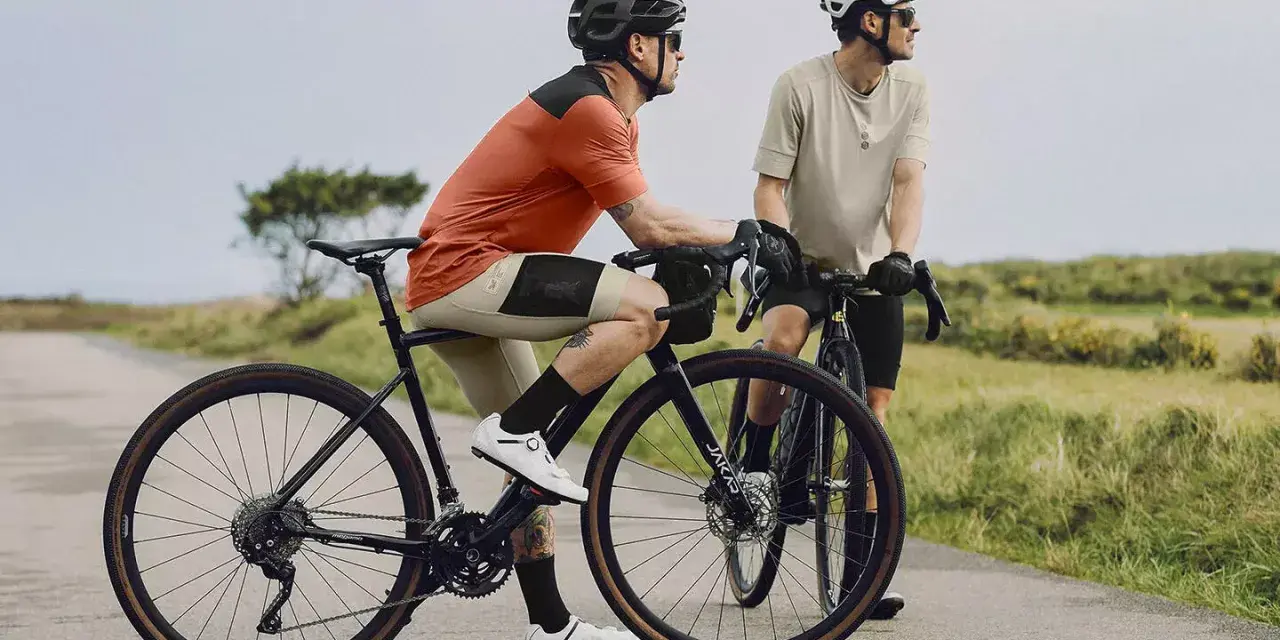 Dwóch rowerzystów na szosie, jeden w pomarańczowej koszulce, drugi w beżowej. To idealny widok na to, co to gravel - wszechstronność i przygoda.