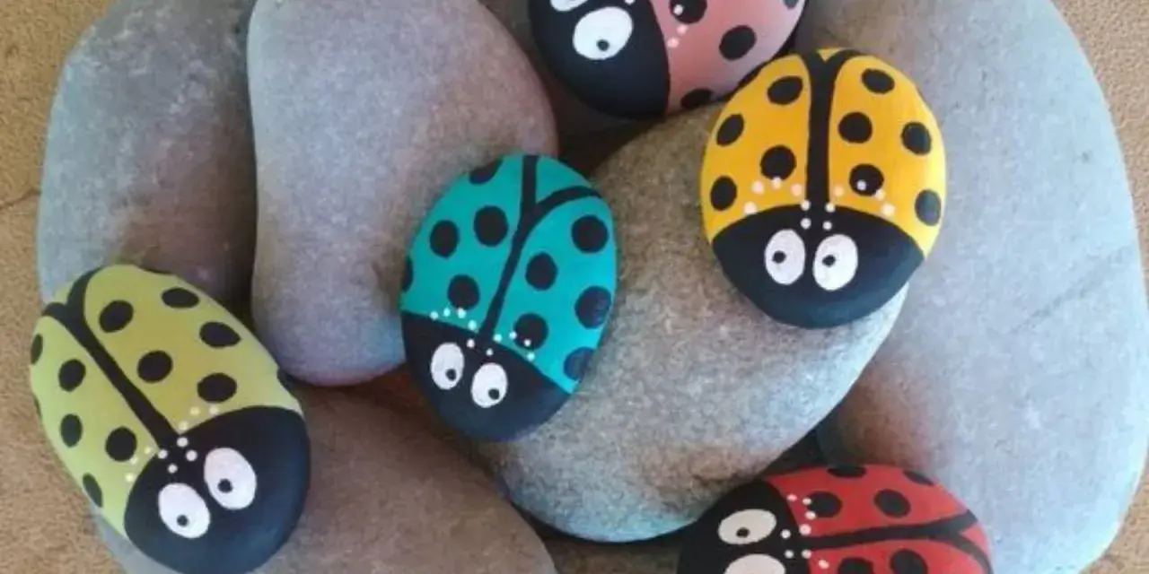 Seis piedras pintadas fáciles con mariquitas de colores: roja, amarilla, verde, rosa y turquesa, descansan sobre piedras grises.