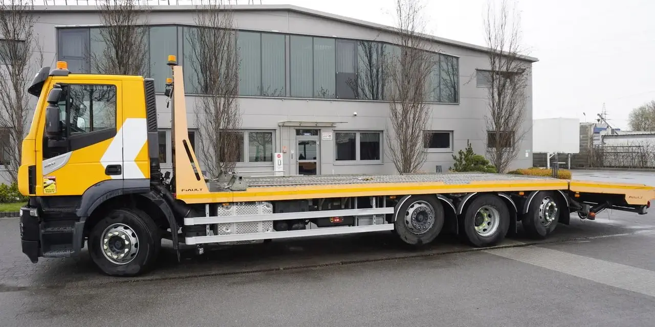 Żółty Iveco Stralis z platformą transportową stoi przed budynkiem.