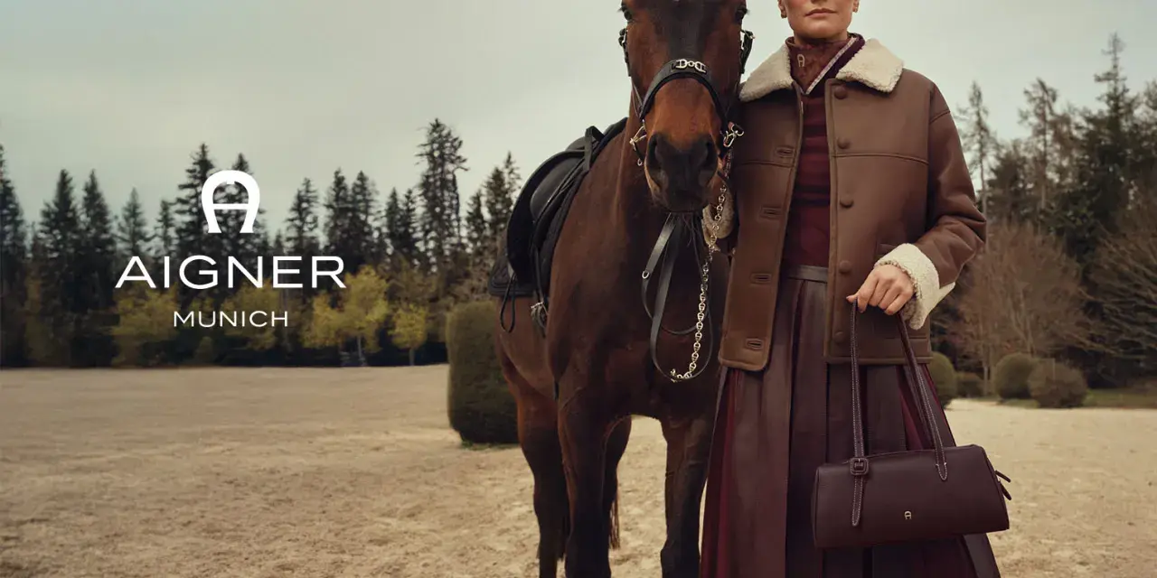 Elegancka kobieta w brązowej kurtce i spódnicy obok konia. Logo AIGNER MUNICH. Aigner co to za marka? Luksusowa moda.
