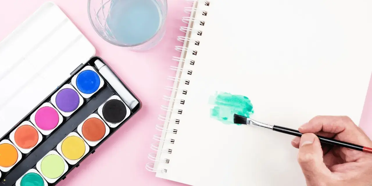 Une main tient un pinceau pour créer une tache verte sur un carnet, à côté d'une palette d'aquarelle et d'un verre d'eau.