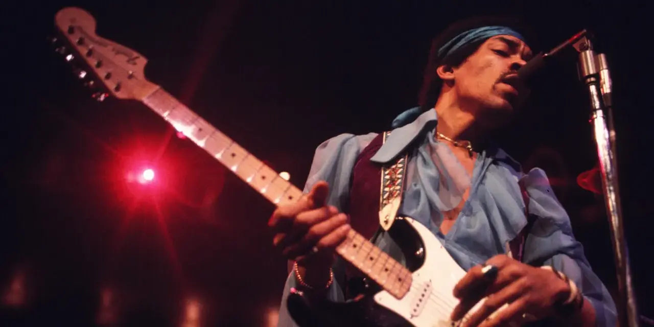 Jimi Hendrix, einer der besten Gitarristen, spielt auf der Bühne seine weiße Fender Stratocaster. Rotes Licht beleuchtet die Szene.