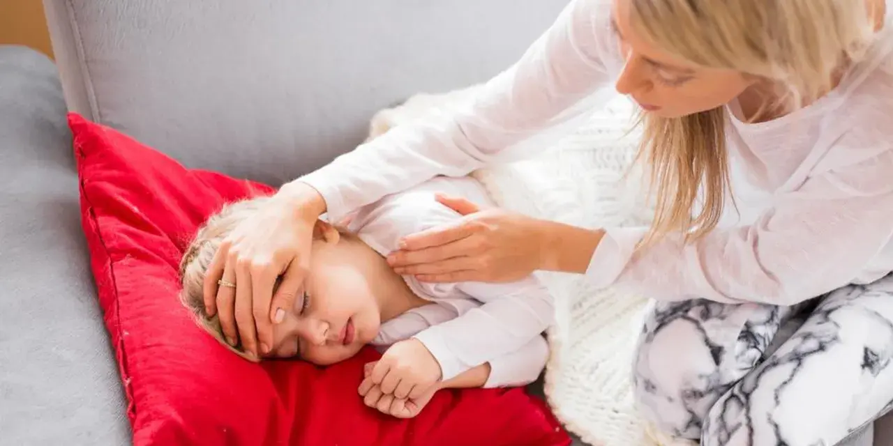Matka troskliwie opiekuje się dzieckiem, które leży na czerwonej poduszce. Dziecko ma zamknięte oczy, a matka delikatnie dotyka jego czoła, sprawdzając, czy nie ma gorączki. To obrazek pokazujący troskę o dziecko podczas jelitówki.