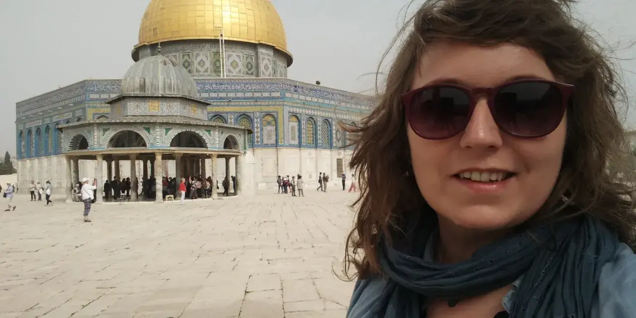 Uśmiechnięta kobieta w okularach przeciwsłonecznych robi selfie na tle Kopuły na Skale. Wokół widać turystów, co sugeruje, że w Jerozolimie jest bezpiecznie.