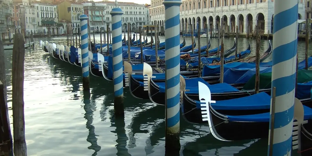 Una fila di imbarcazioni tipiche veneziane, le gondole, ormeggiate lungo un canale, con i loro caratteristici pali a strisce blu e bianche.