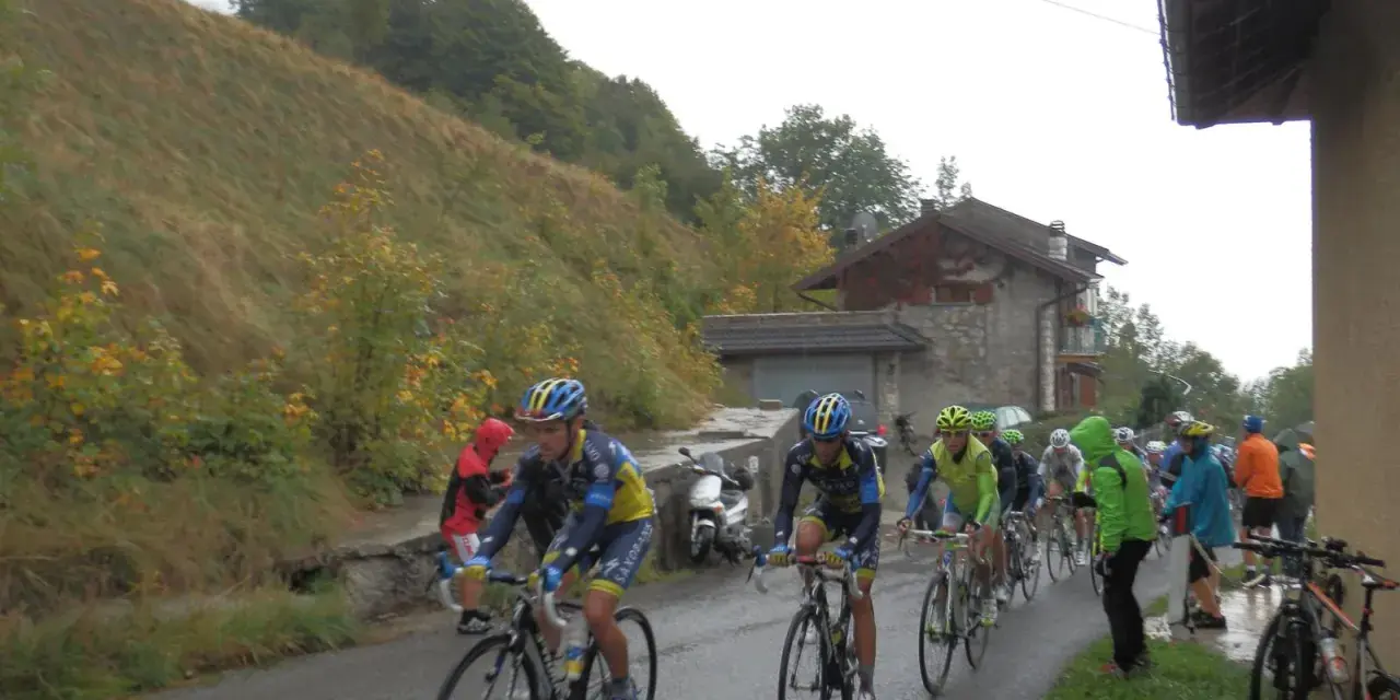 Ciclisti in gara su una strada bagnata, con una **forcella alta** che indica la salita. Il paesaggio è autunnale, con alberi e un pendio erboso.