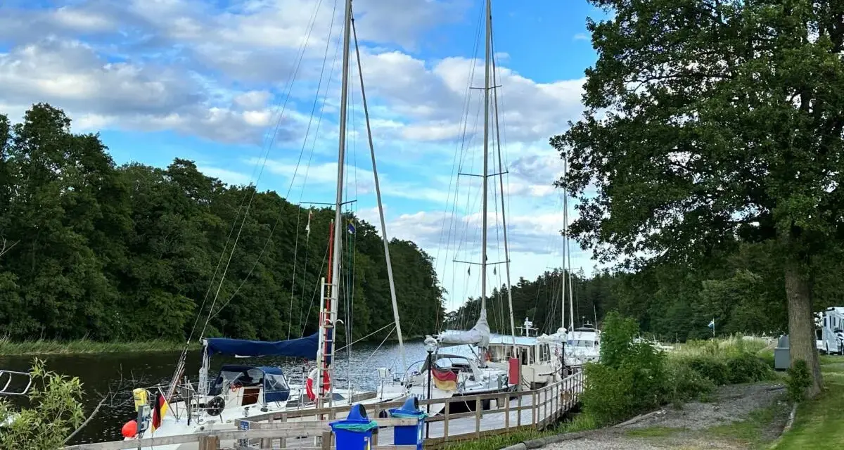 Segelbåtar vid kajen på Dalbergså camping. Grönskande träd och en blå himmel med moln.