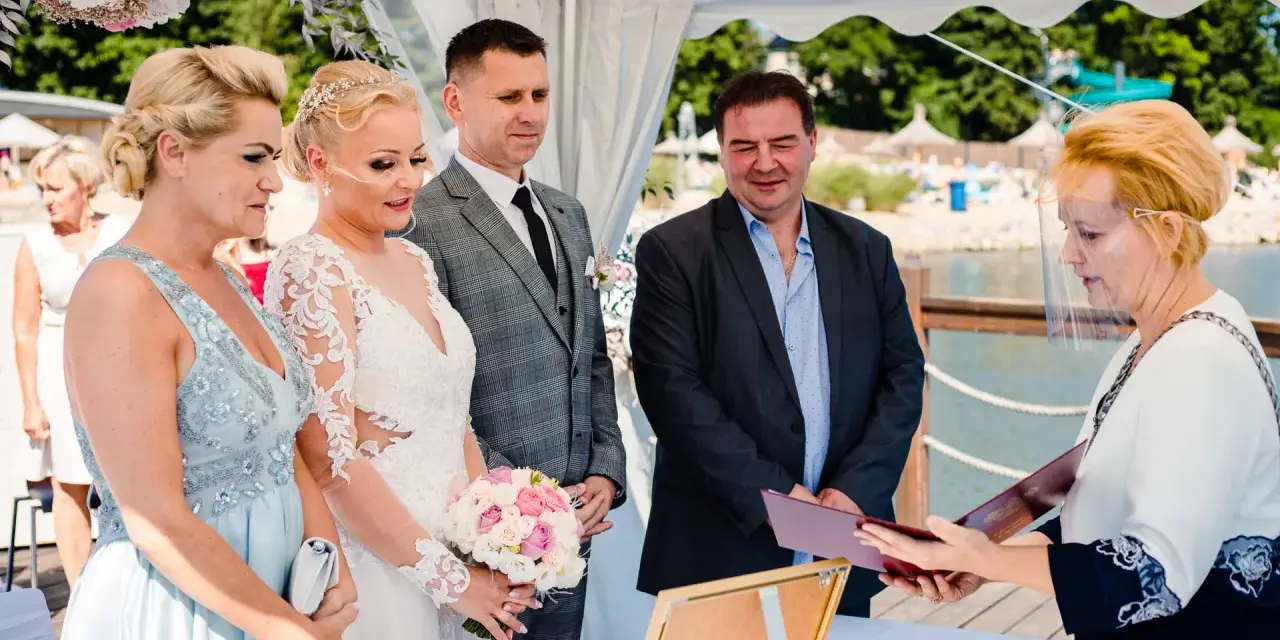 Ceremonia ślubna nad jeziorem, panna młoda w białej sukni, pan młody w garniturze, urzędniczka w przyłbicy.