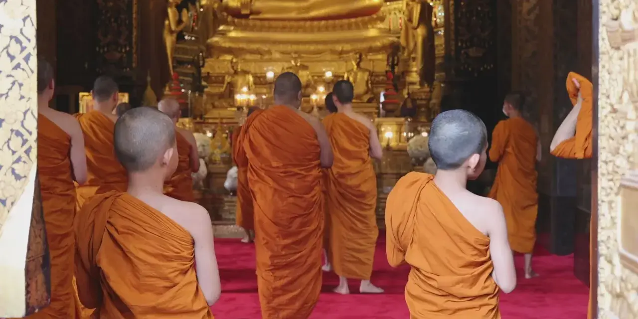 A group of Buddhist monks, another word for ascetics, walk towards a large golden Buddha statue in a temple.