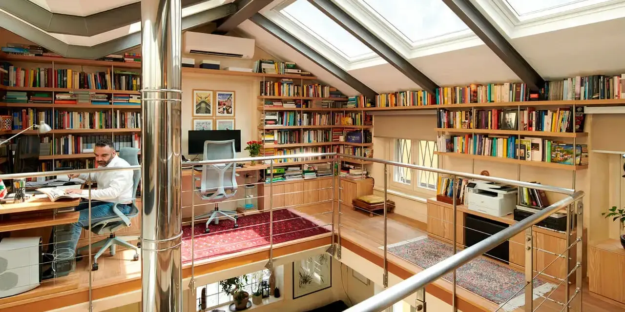 Un uomo lavora in una luminosa libreria mansarda, circondato da scaffali pieni di libri e illuminato da lucernari.