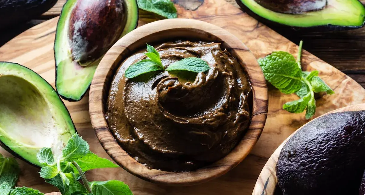 Crema di avocado vellutata in ciotola di legno, circondata da avocado freschi e foglie di menta.