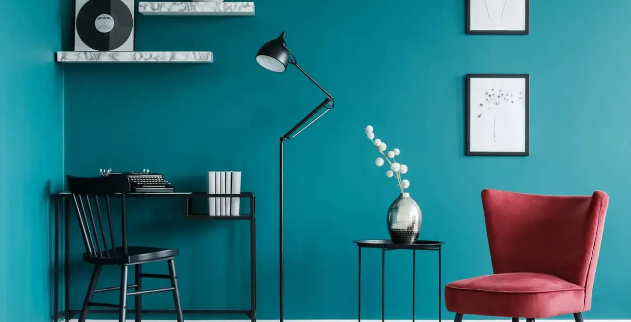 Bureau noir avec machine à écrire, lampe sur pied, fauteuil rouge et étagères sur un mur bleu ou vert canard.