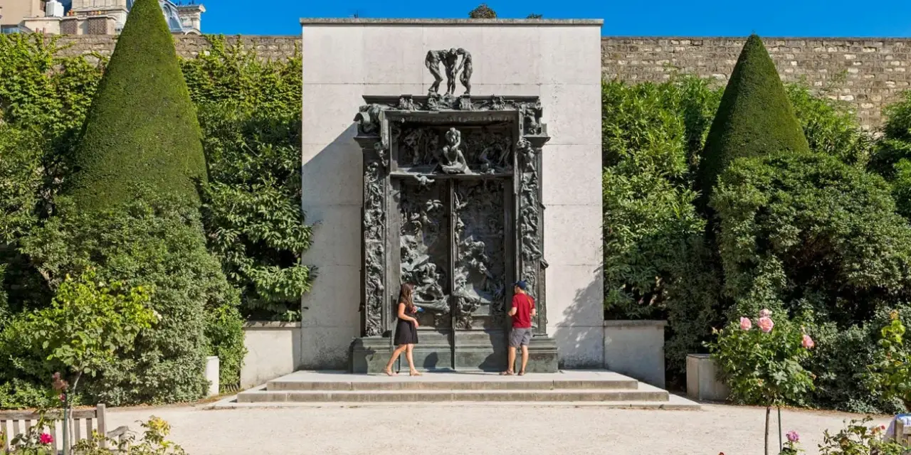 La Porte de l'Enfer, une œuvre monumentale de Rodin, se dresse majestueusement, encadrée par des arbres taillés et un mur de pierre. Deux visiteurs contemplent cette sculpture complexe.