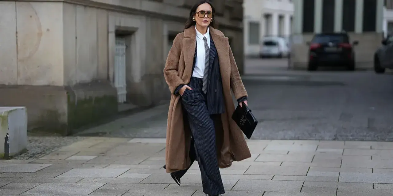 Mujer con abrigo largo marrón, traje de raya diplomática y corbata, luciendo un impecable tomboy style.