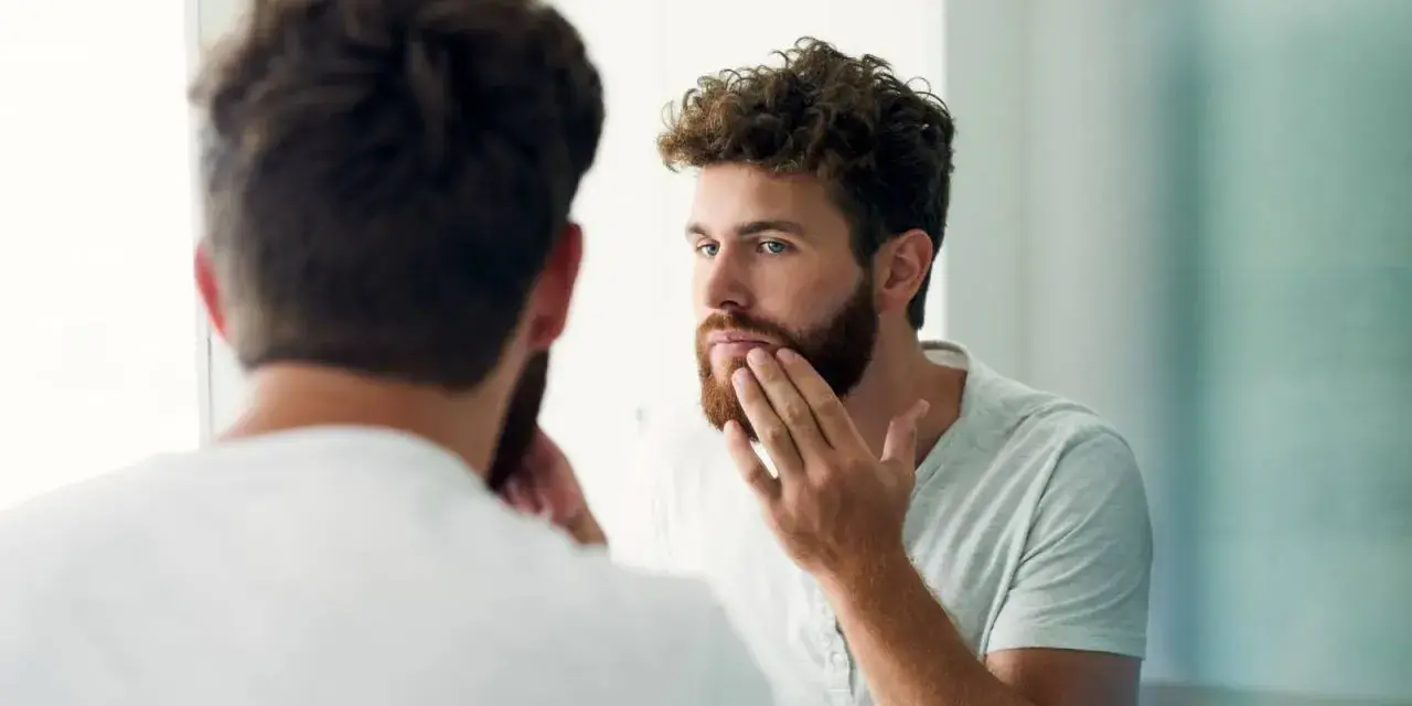 Hombre con barba regia se mira al espejo, acariciando su rostro.