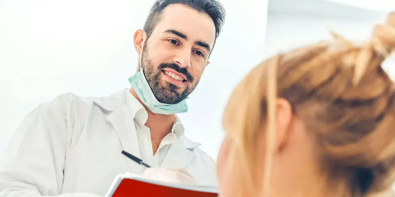 Uśmiechnięty dentysta w białym fartuchu i masce na szyi rozmawia z pacjentką, trzymając notatnik.