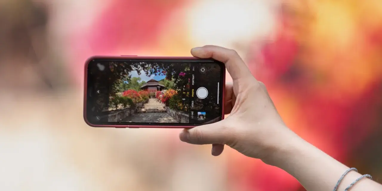 Une main tient un smartphone pour capturer un escalier fleuri menant à un pavillon. L'écran montre les bases de la photographie, avec des options comme "PHOTO" et "PORTRAIT".