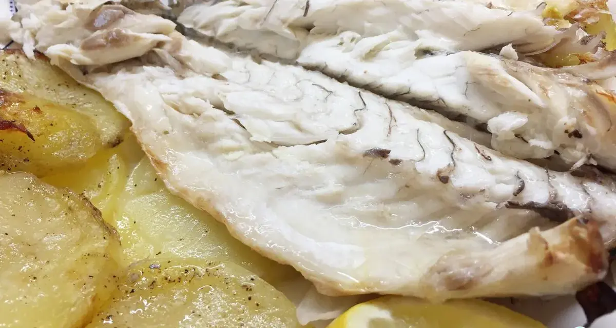 Lubina al horno jugosa y tierna, acompañada de patatas asadas y rodajas de limón fresco.