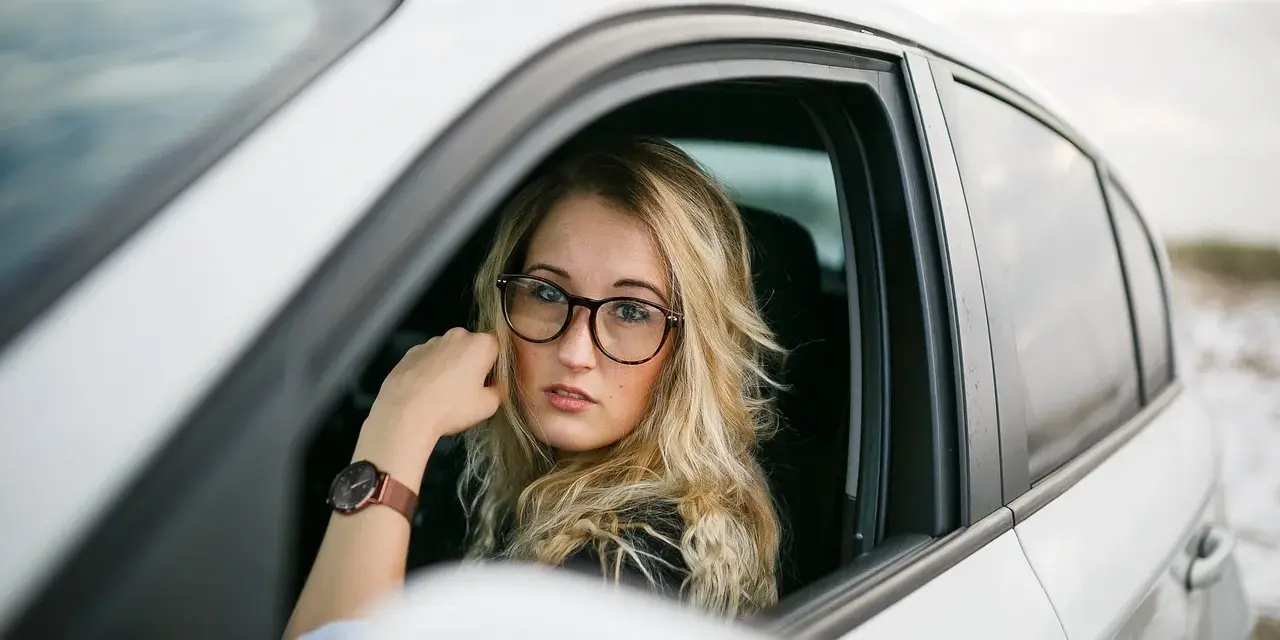 Blondynka w okularach siedzi za kierownicą białego samochodu. Pokazuje, jak nosić okulary, by wyglądać stylowo i pewnie.