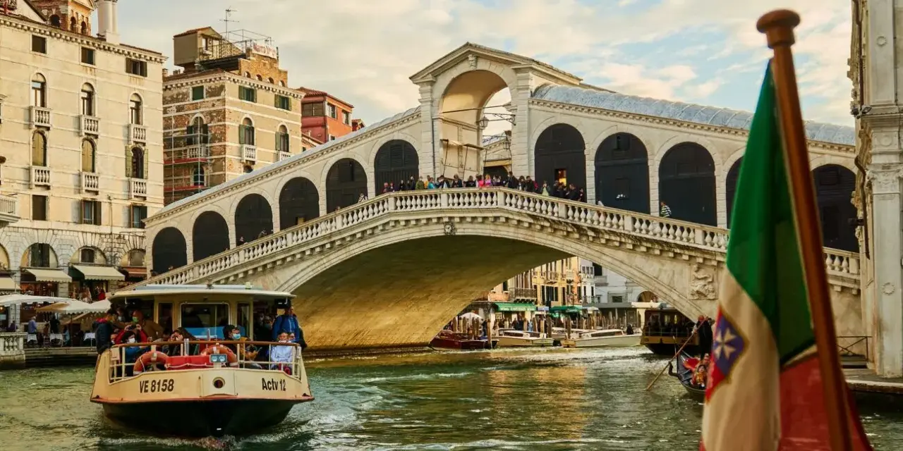 Un vaporetto naviga sul Canal Grande, passando sotto il Ponte di Rialto. L'Italia è rappresentata da una bandiera sventolante.