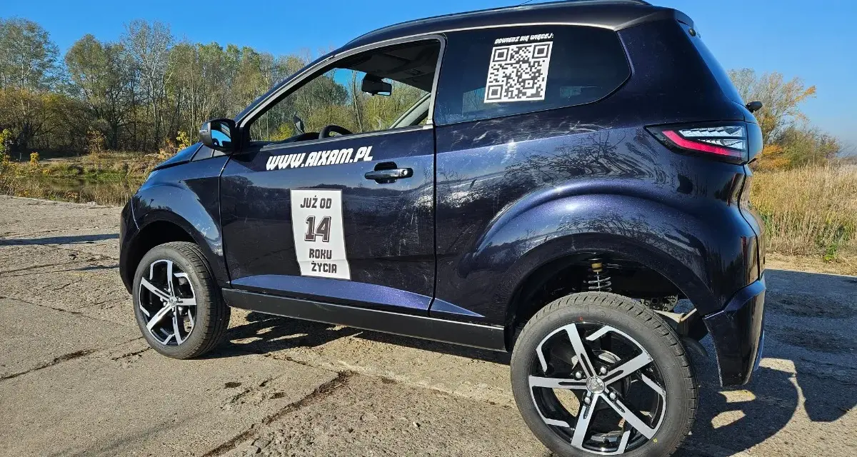 Ciemnoniebieskie Aixam auto z naklejkami "JUŻ OD 14 ROKU ŻYCIA" i kodem QR stoi na tle jesiennego krajobrazu.