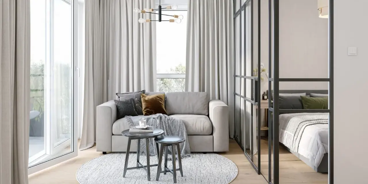 Jasna, nowoczesna kawalerka z wydzieloną sypialnią za szklaną ścianką. W salonie sofa, stoliki i okrągły dywan.
