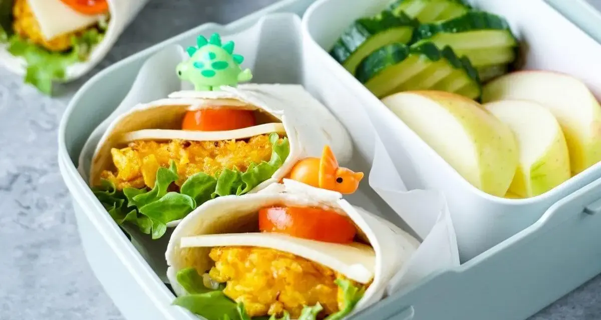 Leckeres Fingerfood für Kinder: Wraps mit Gemüse und Käse, garniert mit kleinen Dino-Spießen. Dazu Gurken und Apfelschnitze.