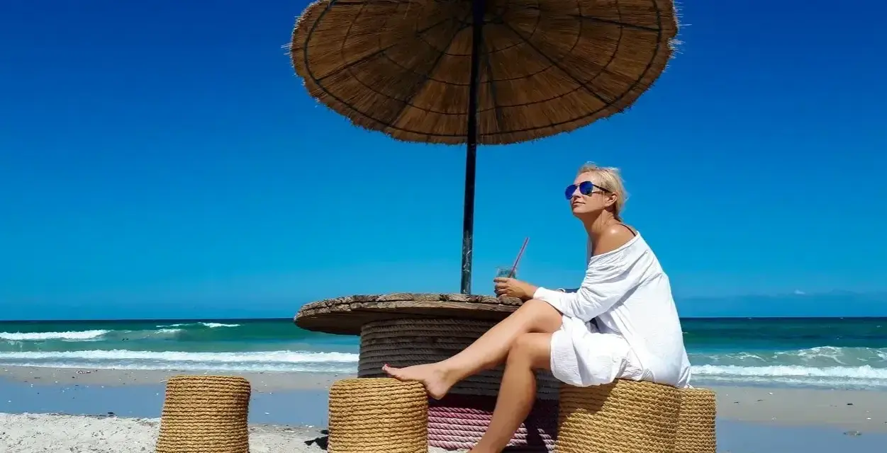 Kobieta w białej sukience relaksuje się pod parasolem na plaży, popijając napój. Idealne tunezja wakacje.