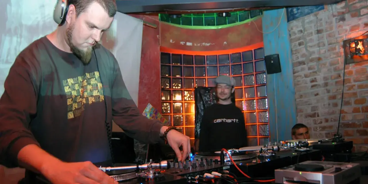 DJ Maceo Wyro w akcji, miksuje muzykę w klubie. W tle widać innych DJ-ów i kolorowe oświetlenie.