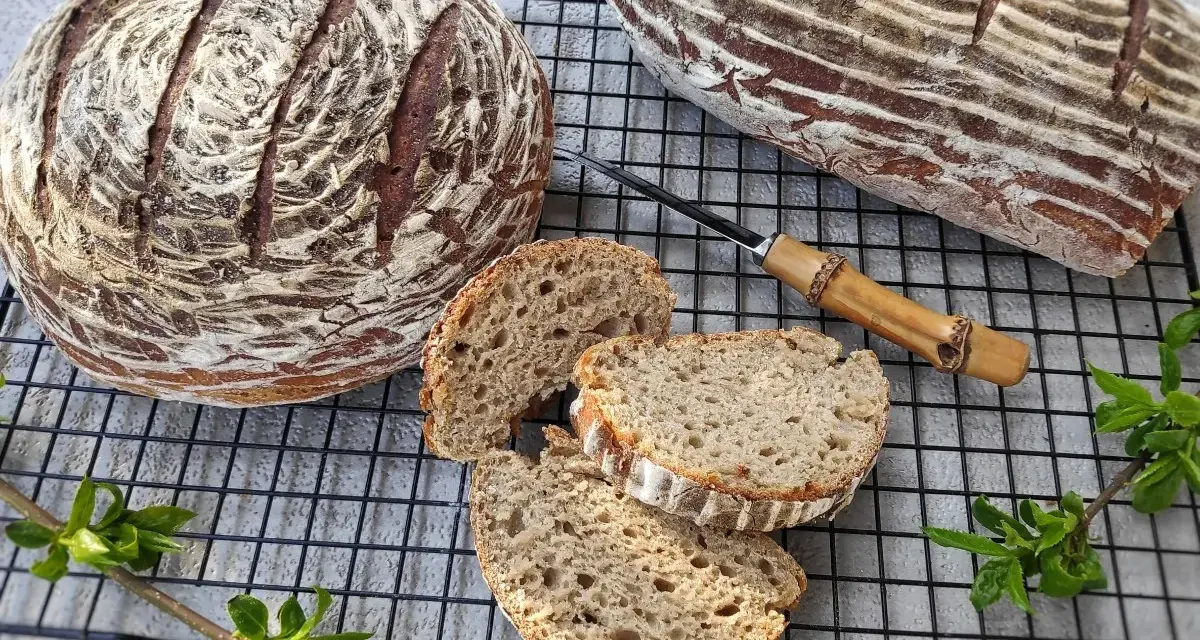 Dwa bochenki chleba żytniego, jeden okrągły, drugi podłużny, leżą na kratce. Obok pokrojone kromki, nóż z drewnianą rączką i zielone gałązki.