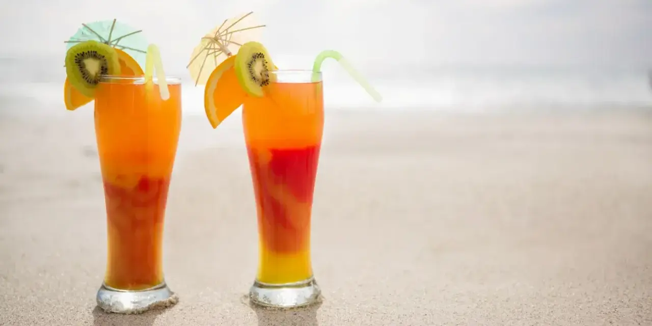 Dwa drinki Malibu z sokiem pomarańczowym, ozdobione plasterkami kiwi i pomarańczy, stoją na piaszczystej plaży.