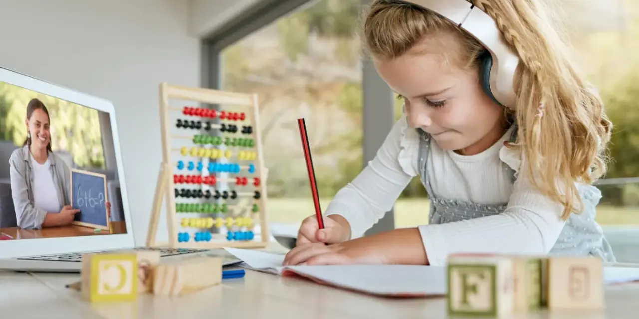 Dziewczynka w słuchawkach uczy się matematyki online, pisząc w zeszycie. Na ekranie laptopa nauczycielka pokazuje zadanie.