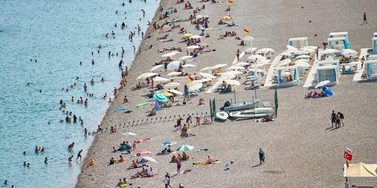 Tłum ludzi odpoczywa na kamienistej plaży w Finike. Niektórzy kąpią się w morzu, inni opalają się pod parasolami.