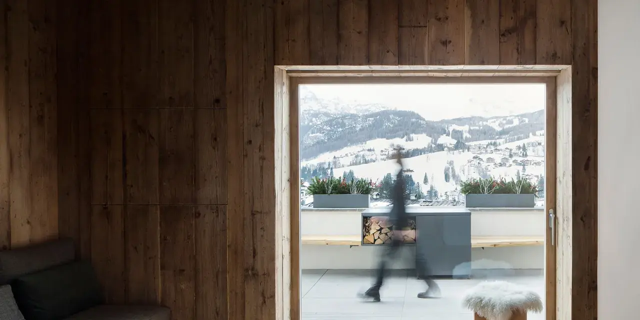 Interni di case di montagna con pareti in legno scuro, travi a vista e un divano grigio. Fuori, un paesaggio innevato.