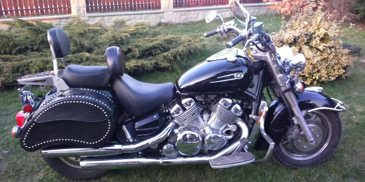 Czarny motocykl Yamaha Royal Star z chromowanymi detalami i skórzanymi sakwami, zaparkowany na trawie przed płotem.
