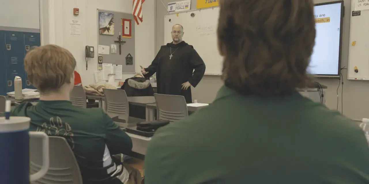 A monastery leader in black robes addresses students in a classroom.