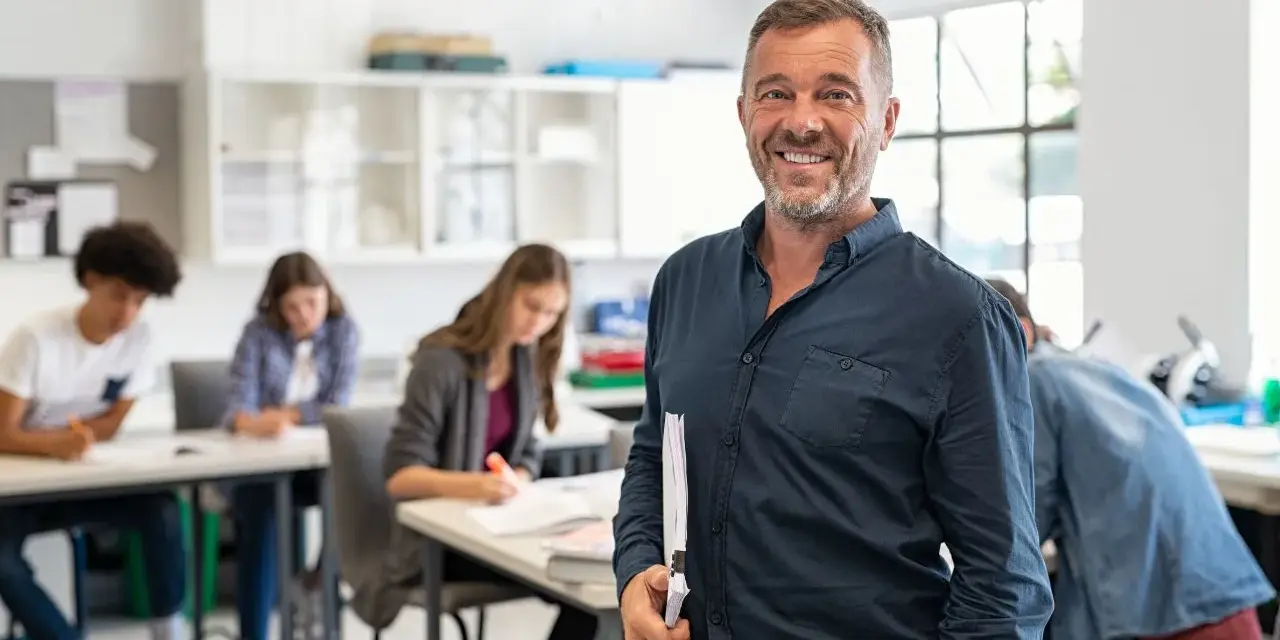 Professore sorridente in aula con studenti. La sua laurea per insegnare alle superiori è evidente dalla sua sicurezza.