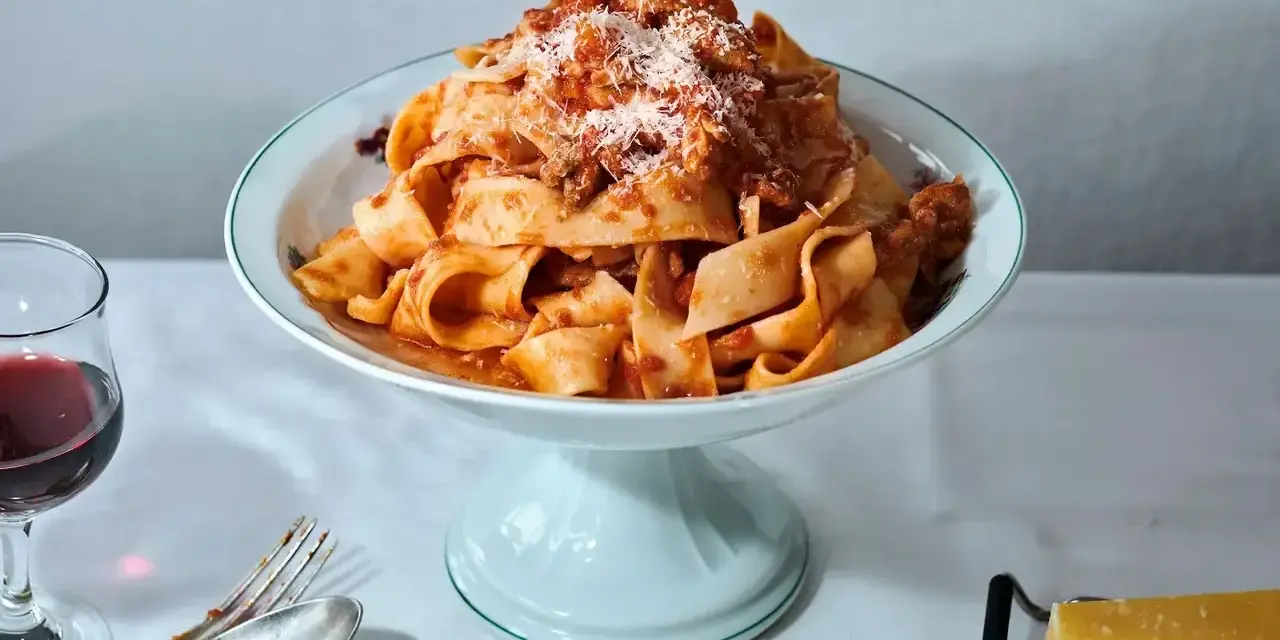 Breite Bandnudeln mit herzhaftem bolognese ragu, bestreut mit Parmesan. Daneben ein Glas Rotwein und Besteck.