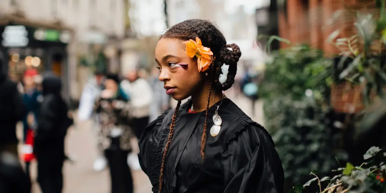 Ragazza con trecce davanti e capelli sciolti, ornata da un fiore arancione e makeup audace. Indossa un abito nero con dettagli voluminosi.