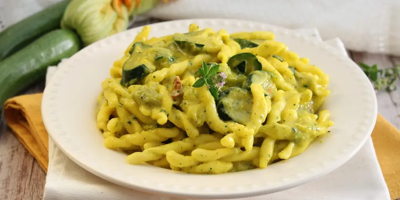 Piatto di pasta con zucchine e zafferano, guarnito con un rametto di erbe aromatiche. Sullo sfondo zucchine fresche e fiori di zucca.